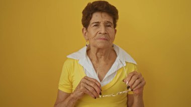 Kısa saçlı, gözlüklü ve sarı gömlekli yaşlı İspanyol kadın, izole edilmiş sarı arka plan duvarına karşı poz verdi..