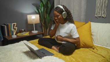 Güzel, kıvırcık saçlı Afro-Amerikan bir kadın, yatak odasında bir yatakta oturuyor, dizüstü bilgisayarın keyfini çıkarıyor, kulaklık takıyor, ve rahat dekor ve kitaplarla çevrili..