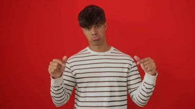 Çizgili gömlekli genç bir İspanyol, izole edilmiş kırmızı bir arka planda duruyor. Üzücü bir ifade sergiliyor. İki başparmağı da aşağıya doğru işaret ediyor..