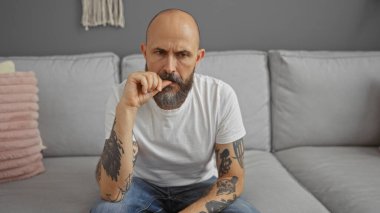 İspanyol bir adam oturma odasında oturuyor, kollarında dövmeleri var, beyaz bir tişört ve kot pantolon giyerken derin düşünceler içinde beliriyor, modern bir ev içinde..