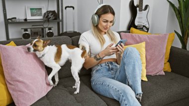 Rahatlamış genç bir kadın kulaklıkla müzik dinler ve köpeğiyle birlikte kapalı bir kanepede akıllı telefonuna göz atar..