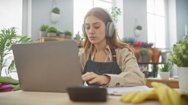 İyi aydınlatılmış bir masada dizüstü bilgisayar kullanan odaklanmış genç bir kadın, kulaklıklarla, modern bir ev ortamında uzaktan kumandalı çalışmalar sergiliyor..