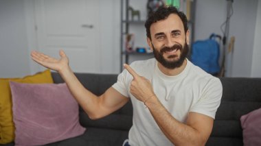 Sakallı, gülümseyen bir adam modern bir oturma odasında bir şey sunuyor.