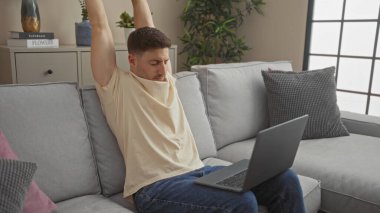 Oturma odasındaki kanepeye dizüstü bilgisayarıyla uzanan genç bir İspanyol, rahat bir ev ortamı sunuyor..
