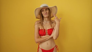 Kırmızı bikinili ve güneş şapkalı güzel bir kadın, izole edilmiş sarı arka planda poz verirken eliyle atış jesti yapıyor..