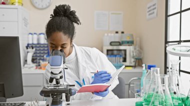 Mikroskop ve not defteriyle laboratuvarda numuneleri analiz eden Afrikalı Amerikalı kadın bilim adamı..