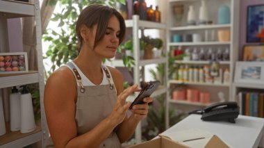 Ev dekorasyonu mağazasında akıllı telefon kullanan kadın çeşitli dekoratif eşyalar ve yeşilliklerle çevrili.