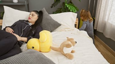 Kızı rahat, aydınlık bir yatak odasındaki pencerenin yanında oynarken bir kadın yatakta dinleniyor..