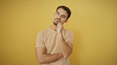 Sakallı, çizgili tişörtlü, sarı arka planda poz veren düşünceli genç bir İspanyol..