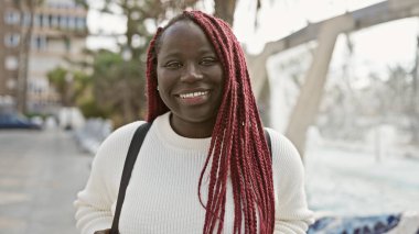 Kırmızı örgülü gülümseyen Afrikalı kadın şehir parkında beyaz kazak giyiyor..