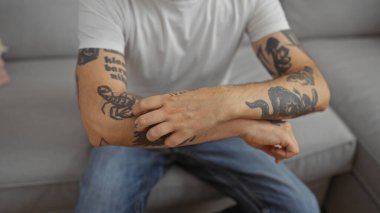 Oturma odasında otururken kolunu kaşıyan dövmeli adam.