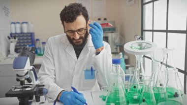 Sakallı bilim adamı örnekleri analiz ediyor ve etrafı cam ve ekipmanlarla çevrili bir laboratuvar ortamında notlar alıyor..