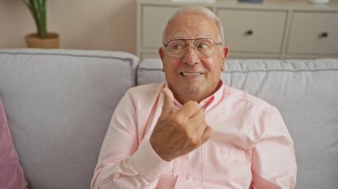 Son sınıf, beyaz, kır saçlı, baş parmağını kaldıran ve rahat bir oturma odasında kanepe ve iç mekan bitkileriyle gülümseyen bir adam..