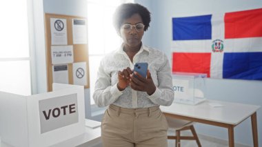 Seçmen kolej odasında akıllı telefon kullanan Afrikalı Amerikalı bir kadın. Arka planda Dominik Cumhuriyeti bayrağı ve oy kullanma kabinleri var..