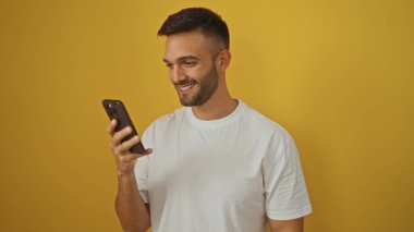 Zeki telefonuna bakarken gülümseyen yakışıklı İspanyol bir adam, canlı sarı bir arka planda izole edilmiş..