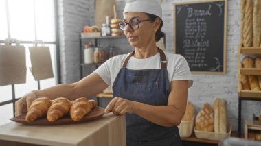 Olgun bir kadın pastanede çalışıyor taze pişmiş kruvasanları arka planda ekmek rafları olan ahşap bir tepsiye yerleştiriyor.