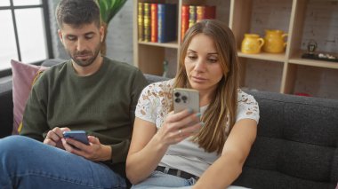 Modern bir oturma odasında akıllı telefonlarla meşgul olan bir çift, çağdaş bir ilişkide kopukluk taslıyorlar..