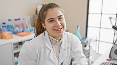 Laboratuvar önlüklü gülümseyen genç bir kadın, etrafı ekipmanlarla çevrili bir tıbbi laboratuvarda çalışıyor..