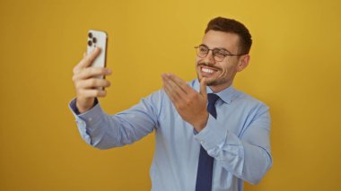 Sakallı, gözlüklü ve mavi gömlekli yakışıklı genç adam, gülümsüyor ve izole edilmiş sarı arka planda selfie çekiyor..