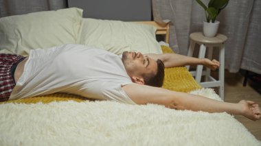 Sakallı genç İspanyol bir adam rahat bir yatak odasında bir yatakta uzanıyor, kollarını içeride yumuşak bir battaniyeye uzatıyor..