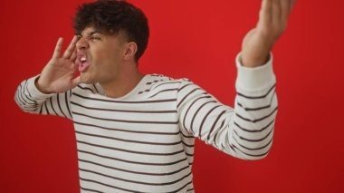 Genç İspanyol bir adam tutkuyla çizgili bir gömlek içinde bağırıyor, canlı kırmızı bir arka plan duvarına karşı izole edilmiş..