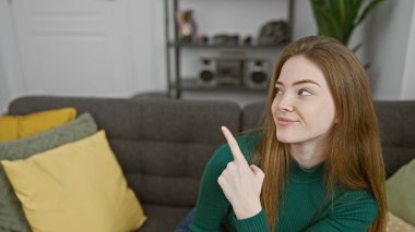 A thoughtful young woman pointing upwards, sitting comfortably in a cozy living room with modern decor. clipart