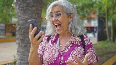 Gri saçlı olgun bir kadın, şehir parkında dikilirken cep telefonuna şaşkın şaşkın bakıyor, canlı bir şehir ortamında açık havada yakalanmış..