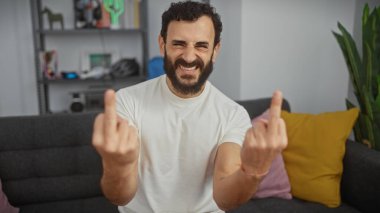 Orta yaşlı, sakallı bir adam sıradan bir oturma odasında kaba bir jest yapıyor..