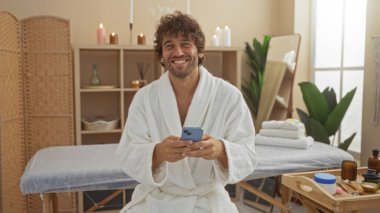 Beyaz cüppeli genç adam elinde bir telefonla kaplıcada oturuyor ve gülümsüyor. Etrafı mumlar, bitkiler ve sağlık dekorlarıyla çevrili. Huzurlu ve davetkar bir ortam yaratıyor..