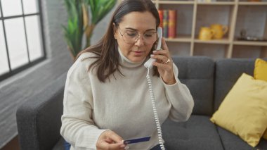 Orta yaşlı bir kadın, elinde kredi kartıyla telefonda konuşuyor. Oturma odasındaki kanepede oturuyor..