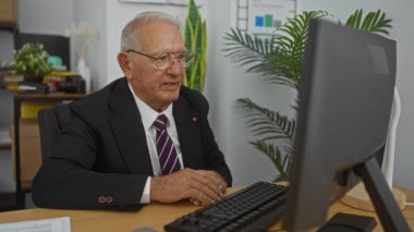 Kapalı bir ofiste bilgisayar başında çalışan kıdemli beyaz bir adam profesyonel bir iş ortamında odak ve deneyim gösteriyor..