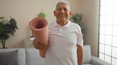 Yakışıklı, gri saçlı İspanyol bir adam evinin rahat oturma odasında yoga minderi tutuyor ve olgun bir yetişkinin aktif yaşam tarzını içeride sergiliyor..