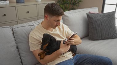 Modern bir oturma odasında daksund tutarken akıllı telefon kullanan genç bir İspanyol, rahat bir kanepeye oturmuş, rahat ve rahat bir atmosfer yaratıyor..