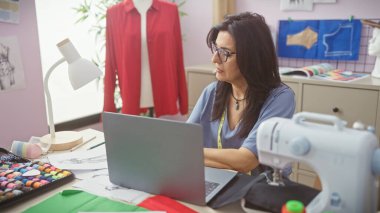 Bir terzide dizüstü bilgisayarda çalışan İspanyol kadın. Etrafı dikiş malzemeleri ve renkli kumaşlarla çevrili..