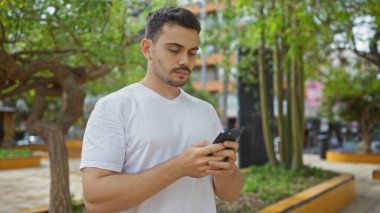 Beyaz tişörtlü yakışıklı bir İspanyol, arka planda binalar ve yeşillikler olan bir parkta dururken akıllı telefon kullanıyor..