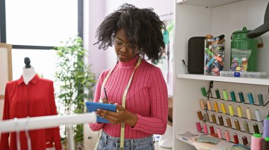 Bir terzi dükkanında renkli iplikler ve bir mankenle çevrili bir tablet kullanan Afrikalı kadın..