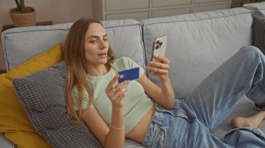 Genç ve çekici sarışın bir kadın, oturma odasındaki bir kanepede uzanmış elinde kredi kartıyla akıllı bir telefona bakarken rahatlamış bir alışveriş anını ya da internet bankacılığı anını yakalıyor..