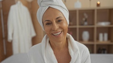 Orta yaşlı İspanyol bir kadın spa odasında beyaz bir elbise ve havlu giyiyor, sıcak bir şekilde gülümsüyor, evde huzur ve rahatlık sağlıyor..