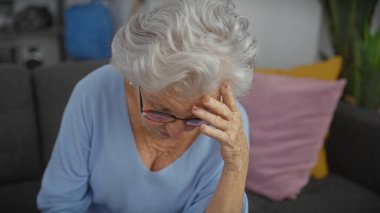 Gri saçlı yaşlı kadın rahat bir oturma odasında stresli hissediyor.