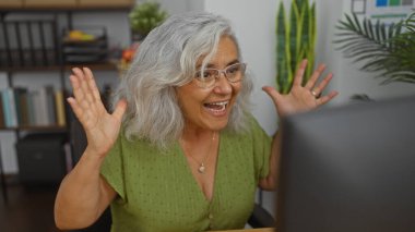 Ofisteki olgun, gri saçlı kadın, bitkiler ve raflarla çevrili bir bilgisayar ekranına heyecanlı bir şekilde bakar ve kapalı alanda neşeli bir tepki gösterir..