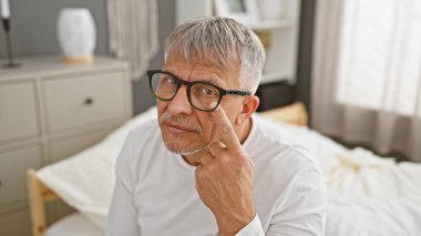 Düşünceli, gri saçlı, gözlüklü bir adam yatak odasında oturur ve rahat ev ortamında bir olgunluk ve tefekkür hissi uyandırır..