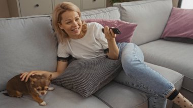 Sarışın genç bir kadın oturma odasındaki kanepeye uzanır. Yanında evcil bir chihuahua 'sı, gülümser ve uzaktan kumandayı tutar. Kapalı bir ortam yaratır..