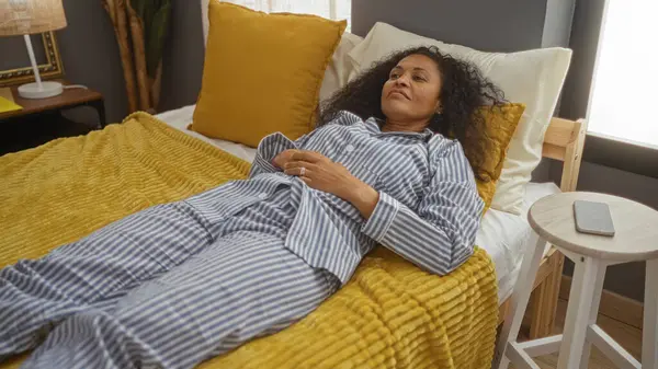 Yatak odasındaki sarı bir yatakta pijamalarıyla dinlenen bir kadın sakin ve rahat bir ev ortamını işaret ediyor.