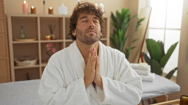 Young man in a white robe meditating in a serene spa room with candles and plants, exuding relaxation and wellness.