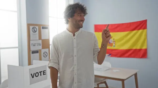 stock image Young man pointing at something inside a spanish electoral college, with a voting booth and a spanish flag in the background.