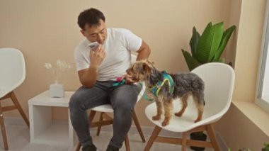 Veteriner kliniğinde oturan Asyalı genç bir adam telefonunu kullanırken küçük köpeğiyle birlikte bir sandalyede oturup evde sakin ve özenli bir an yaratıyor..