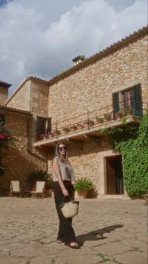 Dışarıda Mallorca İspanya 'da taştan bir evin önünde duran bir kadın güneşli bir günde arkasında çiçekler olan bir hasır çanta tutuyor.