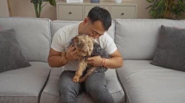 Genç Asyalı bir adam küçük köpeğini kucağına alır ve öper. Gri bir kanepede rahat bir oturma odasında iç mekan bitkileri ve çağdaş dekorlarla otururken..