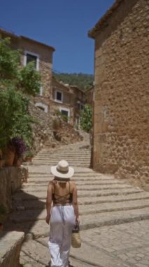 Fornalutx, Mallorca, İspanya 'da bir kaldırım taşı caddesinde tepeye doğru yürüyen bir kadın güneşli bir günde taştan binalar ve yemyeşil alanlarla çevrili güneş şapkası ve beyaz pantolon giyiyor..