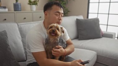 Genç Asyalı bir adam evinde rahat bir oturma odasındaki kanepede evcil köpeğiyle oturuyor, elinde uzaktan kumandayla evde dinleniyor gibi görünüyor..